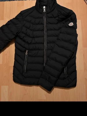 Svart Moncler pufferjacka - Säljer en svart pufferjacka från Moncler med klassisk logga på ärmen och schyssta detaljer som dragkedjor framtill och på fickorna. Insidan är fodrad i vinrött tyg med Moncler-tryck. Jackan har hög krage och är perfekt för kalla dagar.