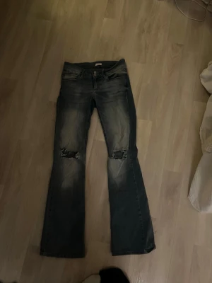 Blå bootcut jeans med slitna knän - Säljer ett par blå bootcut jeans från Nelly i storlek 36. Jeansen har slitningar över knäna och klassiska fickor med lock och knapp bak. Materialet är jeans och färgen är tvättad blå med ljusare partier. Perfekta för dig som gillar en avslappnad och trendig look.