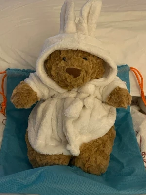 Vit fluffig väst med huva från Jellycat - Supersöt vit fluffig väst från Jellycat med huva och kaninöron. Västen har knytband i midjan och är gjord i mjukt syntetmaterial som känns riktigt gosigt. Perfekt för dig som vill ha något gulligt och mysigt i garderoben.