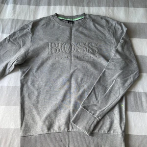 Grå sweatshirt från Hugo Boss - Snygg grå sweatshirt från Hugo Boss med broderad logga framtill. Tröjan har rund halsringning, ribbade muddar och är tillverkad i mjuk bomull. Perfekt för en clean och stilren look.