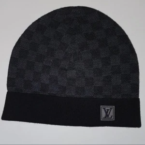 Louis Vuitton mössa  - Snygg svart mössa från Louis Vuitton med diskret rutigt mönster och klassisk LV-logga framtill. Mjuk och skön stickad kvalitet som passar perfekt när det är kallt ute. Stilren design som lyfter din look.