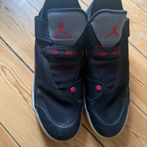 Nike Jordan 33 svart/röd sneakers - US size 14. Säljer ett par Nike Jordan 33 sneakers i svart med röda detaljer och grå plös. Skorna har en modern design med mesh och skinn, samt innovativ snörning med rem och pull-system. Sulan är vit med röda inslag och genomskinlig yttersula. Perfekta för dig som gillar basketstil.