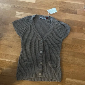 Grå stickad väst med knappar - Snygg grå stickad väst med korta ärmar och v-ringad hals. Västen har fyra stora knappar framtill och två små fickor. Endast använd en gång så lappen sitter inte kvar längre. 100% bomull