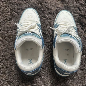 Louis Vuitton blå/vita sneakers - Säljer ett par Louis Vuitton sneakers i vitt och ljusblått med klassiskt LV-monogram på denimdetaljerna. Skorna har snörning, rund tå och platt sula. Materialmix av skinn och denim ger en lyxig känsla och cool kontrast. Perfekta för dig som vill sticka ut med designerstil.