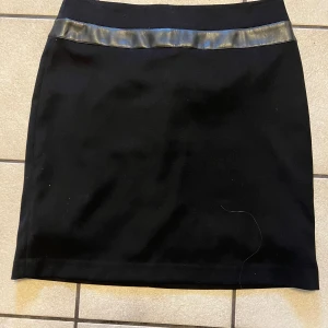 Svart kortkjol från DiKa med skinnband - Snygg svart kortkjol från DiKa med ett stilrent skinnliknande band upptill. Kjolen har en klassisk rak passform och dragkedja bak. Perfekt för dig som gillar en enkel men edgy look.