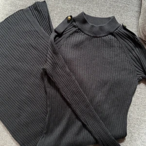 Svart ribbad klänning - Svart ribbad stickad klänning från L.O.G.G. i storlek L. Klänningen har en mjuk och stretchig känsla. Snygga detaljer med ribbad struktur och dekorativa knappar vid axlar. Perfekt för en trendig look.