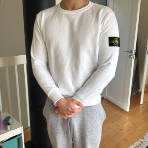 Vit långärmad tröja Stone Island - Vit långärmad tröja från Stone Island med klassisk rund hals och ikonisk svart patch med logga på vänster ärm. Tröjan är i mjuk bomull och har en clean, enkel look som passar perfekt till jeans eller sweatpants. Den passar S mer. Har en exakt samma i grå med skriv vid intresse 