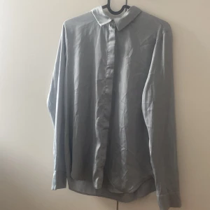Ljusgrå skjorta från H&M, XS - Snygg gråblå skjorta från H&M i storlek XS. Skjortan har klassisk krage, dolda knappar framtill och långa ärmar med manschett. Materialet är mjukt och har en lätt glansig finish, vilket ger en clean och modern look.