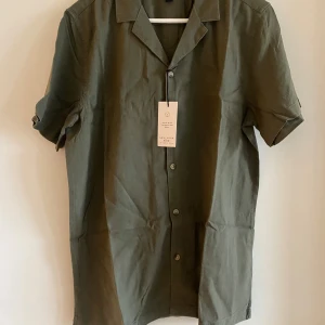 NY Herr linneskjorta S  - Olivgrön skjorta från New Look med klassisk krage och knappar framtill. Tillverkad i en skön mix av linne och viskos som ger en lätt och luftig känsla. Kortärmad modell med avslappnad passform, perfekt för varma dagar. Enkel och stilren enfärgad design Ny med lappar kvar!