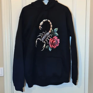 Svart hoodie med skorpion och ros XXL - Svart varm hoodie i storlek XXL med en stor, detaljerad print av en skorpion och en röd ros på framsidan. Tröjan har huva med snörning och en klassisk magficka. Perfekt för dig som gillar edgy och unika motiv. Tyvärr kan man bara ladda upp fyrkantiga bilder här på Plick. Vill du ha bättre bilder så skriv till mig.