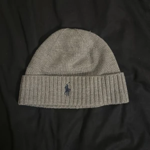 Grå mössa från Polo Ralph Lauren - Snygg grå mössa från Polo Ralph Lauren i 100% merinoull. Klassisk ribbstickad kant och broderad marinblå logga framtill. Perfekt för kalla dagar och enkel att matcha med olika outfits.