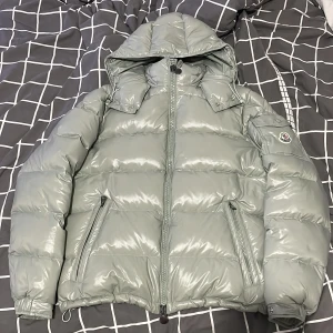 Ljusgrå Moncler pufferjacka med huva - Säljer en sjukt snygg ljusgrå pufferjacka från Moncler. Jackan har glansig finish, stor huva och dragkedja framtill. Två snedställda fickor med dragkedja och klassisk Moncler-logga på ärmen. Perfekt för kalla dagar när du vill vara både varm och stilren. Fick den i present men den passade inte så vill bli av med den skriv dm om ni har frågor.
