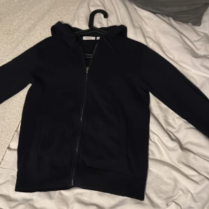 Mörkblå zip - Mörkblå zip från RYVLS med hel dragkedja framtill och huva med snören. Tröjan har två fickor på sidorna och är långärmad. Perfekt för chill dagar eller när du vill ha en enkel och stilren look.