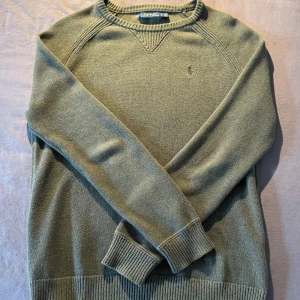 Olivgrön stickad tröja från Polo Ralph Lauren - Unik och snygg olivgrön stickad tröja från Polo Ralph Lauren med diskret broderad logga på bröstet. Tröjan har rund halsringning, ribbade muddar och är långärmad. Perfekt för lager-på-lager under kyliga dagar. Det är bara att höra av dig om du har någon fundering! 