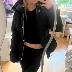 Svart pufferjacka med huva - Säljer en svart pufferjacka med huva och dragkedja. Jackan har en oversized passform och dragsko i midjan för att justera siluetten. Perfekt för kyligare dagar och riktigt skön att bära. Stängs med både dragkedja och knappar framtill. Helt oanvänd så i mycket gott skick - köpt på zalando.