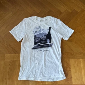 Vit t-shirt Puerto Banús print - Vit t-shirt med tryck av klocka, champagneflaska, yacht och texten 'Puerto Banús' på framsidan. Klassisk rund hals och korta ärmar. Perfekt för dig som gillar chill och lyxiga vibbar. Storlek S