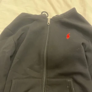Svart hoodie från Polo Ralph Lauren - Svart hoodie från Polo Ralph Lauren med röd broderad logga på bröstet. Tröjan har huva, dragkedja framtill och två fickor. Materialet är mjukt och känns som bomull, perfekt för chill dagar. Klassisk och stilren design som alltid funkar.