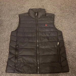 Ralph Lauren väst  - Svart dunväst från Polo Ralph Lauren med horisontella quiltade sömmar och röd logobrodering på bröstet. Västen har hög krage, två sidofickor med dragkedja och en smidig dragkedja framtill. Perfekt för lager-på-lager under kyliga dagar. Storlek 18-20 vilket motsvara ca xs 