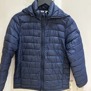 Blå pufferjacka från GAP Kids XL - Blå pufferjacka från GAP Kids i storlek XL. Jackan har huva, horisontella quiltade sömmar och dragkedja framtill. Perfekt för kyliga dagar och har en clean, sportig look. Materialet är syntet och jackan är långärmad. Passar Stl XS 