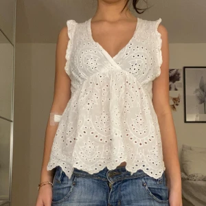 Vit broderad blus med volang - Superfin vit blus med broderade detaljer och volangkant. Blusen har v-ringad omlottfront, ärmlös design med små volanger vid axlarna och ett luftigt, lätt material som känns perfekt för varma dagar. Snyggt mönster med hålbroderi och söt scallopad nederkant.