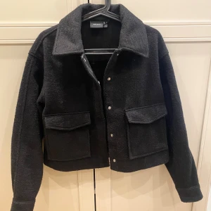 Svart teddyjacka från Vero Moda - Svart jacka från Vero Moda med stora fickor framtill och klassisk krage. Jackan är croppad och har tryckknappar. Perfekt för dig som gillar en stilren och trendig look med lite boxig passform.