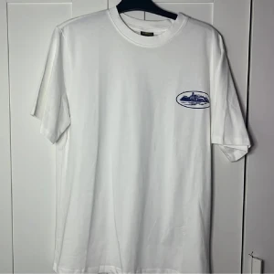 Vit t-shirt med blått tryck - Vit t-shirt med rund hals och korta ärmar. På bröstet finns ett blått tryck med motiv av en byggnad, och på ryggen står det 'RULES THE WORLD' i stort blått tryck. Materialet känns som mjuk bomull, perfekt för en chill och stilren look.