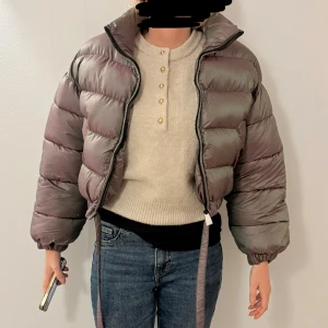 Lila pufferjacka med snörning - Säljer en croppad pufferjacka i metallic-lila med dragkedja och snörning nertill. Jackan har hög krage, långa ärmar. kort vinterjacka, helt ny i strl S säljes för 200kr