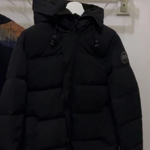 Svart dunjacka från Canada Goose - Säljer min svarta macmiller från Canada Goose med huva och dragsko. Jackan har klassisk patch på ärmen och är riktigt varm och fluffig. Perfekt för kalla dagar och har en clean, stilren look som funkar till allt. Har använt jackan bara under en vinter eftersom den var smal på mig med annars allt e bra/som ny skick.
