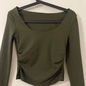 Olivgrön långärmad topp med rynk - Snygg olivgrön långärmad topp med rundad, djup urringning och rynkade detaljer i sidorna. Toppens material känns mjukt och stretchigt, perfekt för en trendig och bekväm look.