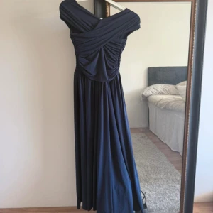 Marinblå långklänning från H&M XS - Elegant marinblå långklänning från H&M i storlek XS. Klänningen har omlott-detaljer upptill, vackert draperad byst och avskuren midja. Materialet är mjukt och följsamt, perfekt för en stilren look. Offshoulder-modell som ger en snygg siluett.