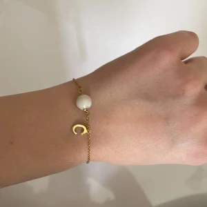 Guldarmband med pärlor och C-berlock - Fint och tunt armband i guldton med två vita pärlor och en liten rund guldplatta. Armbandet har även en berlock i form av bokstaven C. Perfekt för dig som vill ha något personligt och stilrent på handleden.