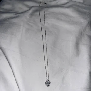 Silverhalsband med hjärta från Pandora - Snyggt halsband i silverfärg med ett hjärtformat hänge dekorerat med små glittrande stenar. Kedjan är tunn och stilren, perfekt för att ge en extra touch till din outfit. Hänget har en detaljrik design som verkligen står ut.