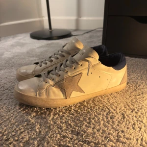 Golden Goose skor - Golden Goose Superstar sneakers i vitt skinn med grå mockastjärna på sidan och mörkblå häl. Skorna har grå snörning, rund tå och platt sula i gummi. Märkets logga syns på sidan och innersulan. Skriv för funderingar/fler bilder!