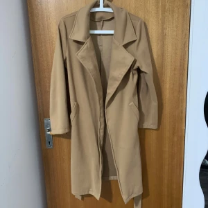 Beige kappa med bälte och krage - Stilren beige kappa med klassisk krage och knytskärp i midjan. Långärmad modell med två fickor framtill och rak passform. Perfekt för dig som vill ha en enkel men snygg look till höst och vår.
