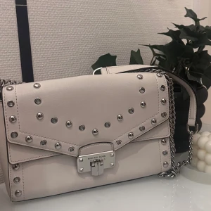 Vit axelväska från Michael Kors - Snygg vit axelväska från Michael Kors i skinn med silverfärgade nitar och kedjedetaljer. Väskan har ett lås framtill med logga och stilren design. Perfekt storlek för det viktigaste och ger en cool touch till din outfit.