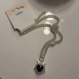 Silverhalsband med svart hjärthänge - Snyggt silverfärgat halsband från Glitter med ett coolt hänge i form av ett svart hjärta omgivet av glittriga detaljer och spetsiga kanter. Kedjan är grov och ger ett trendigt intryck, perfekt för att lyfta din outfit.