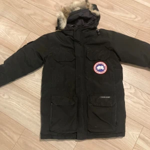 Svart parkas från Canada Goose med päls - Säljer en svart Canada Goose parkas med ikonisk patch på bröstet och huva med päls. Jackan har flera stora fickor framtill och är fodrad med dun och fjädrar för extra värme. Behöver bara en snabbtvätt 