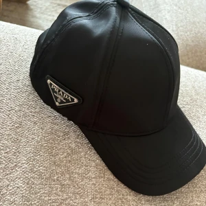 Svart keps från Prada - Snygg svart keps från Prada med klassisk böjd skärm och ikonisk metalltriangel med logga på sidan. Kepsen har stilrena sömmar och är tillverkad i ett slätt syntetmaterial som ger en exklusiv vibe. Perfekt för dig som gillar designeraccessoarer.