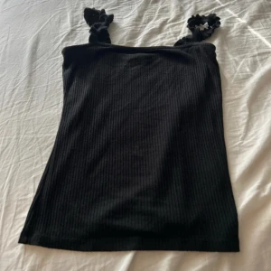 Svart ribbad linnetopp med volangband - Säljer en svart ribbad linnetopp med söta volangprydda axelband. Toppen har en tight passform och är perfekt för dig som gillar en enkel men ändå detaljrik stil. Materialet är mjukt och stretchigt för extra komfort.