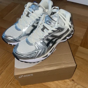 ASICS sneakers i silver och vitt - Snygga ASICS sneakers med coola detaljer i silver och vitt, svarta ränder på sidorna och vit mesh upptill. Skorna har snörning och en chunky platt sula som ger grym komfort. Perfekta för dig som gillar sportig stil och vill sticka ut med metallic-vibe.