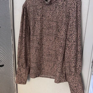 Beige prickig långärmad blus - Säljer en beige blus  med svarta prickar och långa ärmar. Blusen har en lite högre krage med voulang, knappar vid ärmslut och en rak passform. Perfekt för dig som gillar mönster och vill sticka ut lite extra.