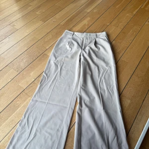 Beige vida kostymbyxor - Snygga beige kostymbyxor med vida ben och klassisk passform. Byxorna har hög midja, bälteshällor och pressveck framtill för en clean look. Perfekta för dig som gillar stilrena och trendiga outfits.