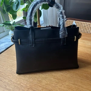 Svart handväska från Mango gulddetaljer - Stilren svart handväska från Mango med guldfärgade spännen och dragkedja. Väskan har två handtag och en klassisk, rektangulär form. Tillverkad i slätt skinnliknande material och har praktiska innerfack med dragkedja. Helt oanvänd och ny med prislapp! Först till kvarn❣️