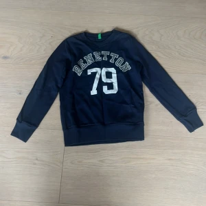Mörkblå sweatshirt från Benetton - Mörkblå sweatshirt från Benetton med tryckt text och siffran 79 i vitt framtill. Klassisk rund halsringning och långa ärmar. Perfekt för en sportig och avslappnad stil.