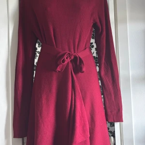 Vinröd omlottklänning med bälte - Säljer en vinröd långklänning med omlott-design och knytbälte i midjan. Klänningen har långärmade ribbade ärmar och en asymmetrisk nederdel som ger en cool och modern look. Materialet är stickat och mjukt, perfekt för dig som gillar att sticka ut med stil.