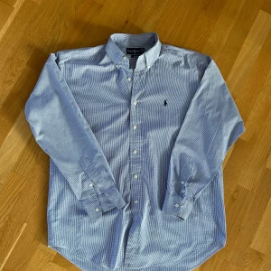 Blårutig skjorta från Ralph Lauren - Snygg blårutig skjorta från Ralph Lauren, modell Blake. Skjortan har klassisk krage, knappar hela vägen och lång ärm med manschett. Materialet är mjuk bomull och den har det ikoniska Polo-logot broderat på bröstet. Perfekt för dig som gillar stilren och tidlös design.