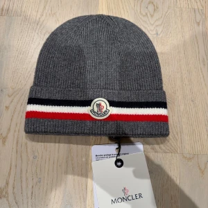 Grå Moncler mössa med ränder - Snygg grå ribbstickad mössa från Moncler med bred uppvikt kant och klassiska Moncler-loggan framtill. Mössan har marinblå, vit och röd rand längs kanten för en sportig vibe. Perfekt för dig som gillar stilrena accessoarer med premiumkänsla.