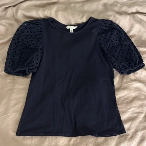 Mörkblå topp med broderade puffärmar H&M - Supersnygg mörkblå topp från H&M med broderade puffärmar i bomull. Ärmarna har ett coolt hålmönster och resår vid avslut. Kroppen är enkel och mjuk, perfekt för en trendig look. Rund halsringning och normal passform.