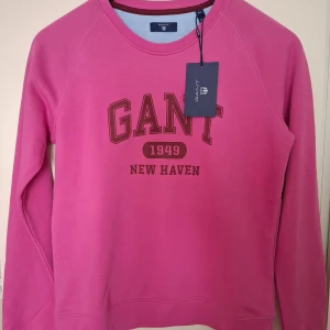 Ny GANT rosa sweatshirt - Snygg rosa sweatshirt från GANT med tryck 'GANT 1949 NEW HAVEN' framtill. Klassisk rund halsringning och långa ärmar. Mjuk bomullskvalitet som är skön att bära. Perfekt för en avslappnad och sportig stil. Helt ny med lapp kvar!
