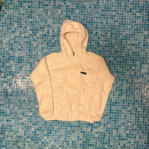 Vit fleecehoodie från Patagonia - Mysig vit hoodie från Patagonia i fluffig fleece med huva och half zip-dragkedja. Tröjan har en relaxed passform, lång ärm och en stor känguruficka framtill. Perfekt för kyliga dagar när du vill vara både varm och stilren.
